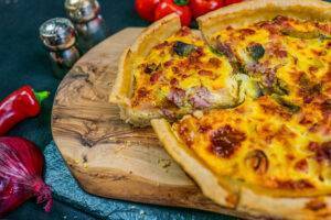 bacon and leek quiche on a wooden chopping board next to onion and chilli